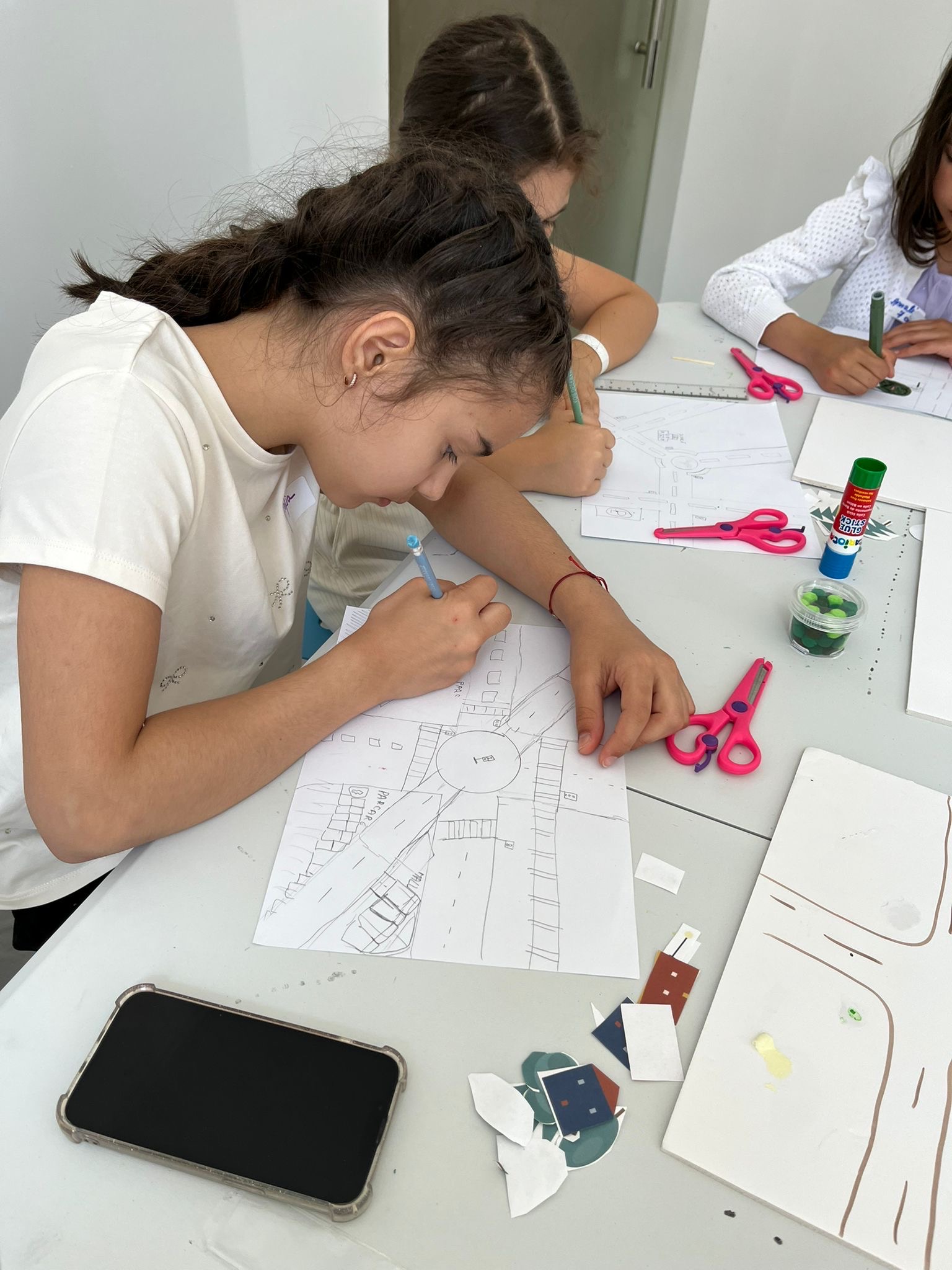 O fetiță schițează în creion un plan de stradă cu sens giratoriu și clădiri, la masa de atelier