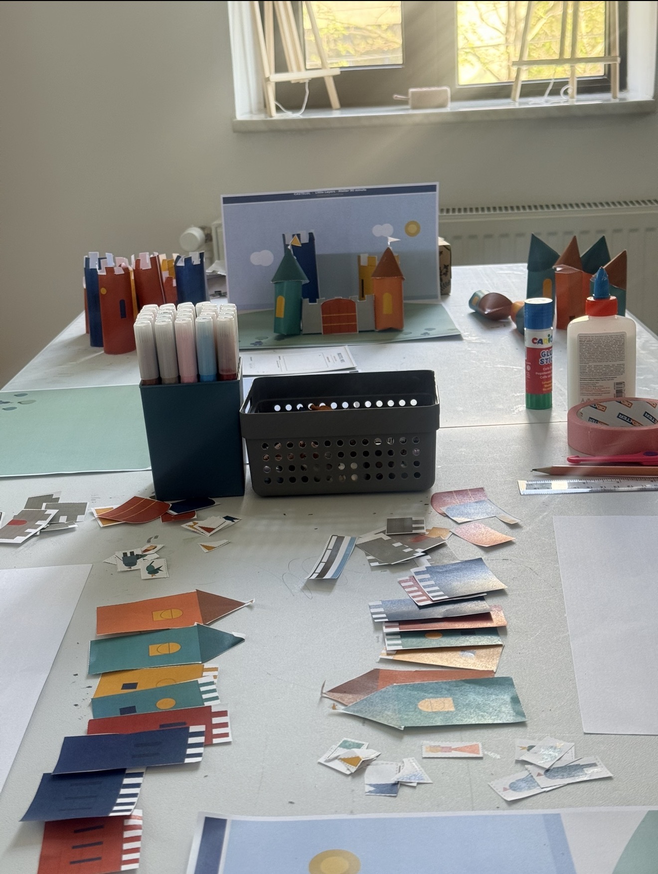 Un castel de hârtie cu turnuri colorate și steaguri, în lumină caldă de fereastră, construit la atelier